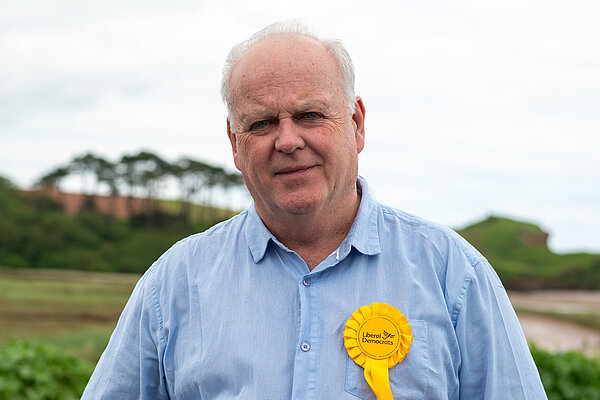 Paul Arnott with a yellow Liberal Democrats rosette against a scenic background with trees and grass