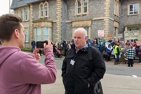 Paul Arnott being filmed by a reporter in front of protestors at Exmouth Town Hall